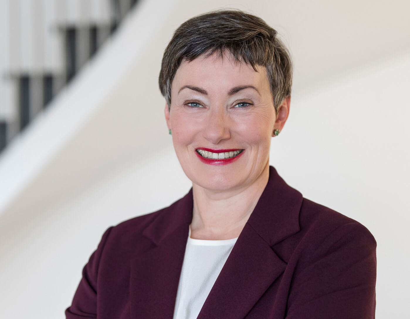 Claudia Ruf, lächelnd im weinroten Blazer vor einer hellen Treppe